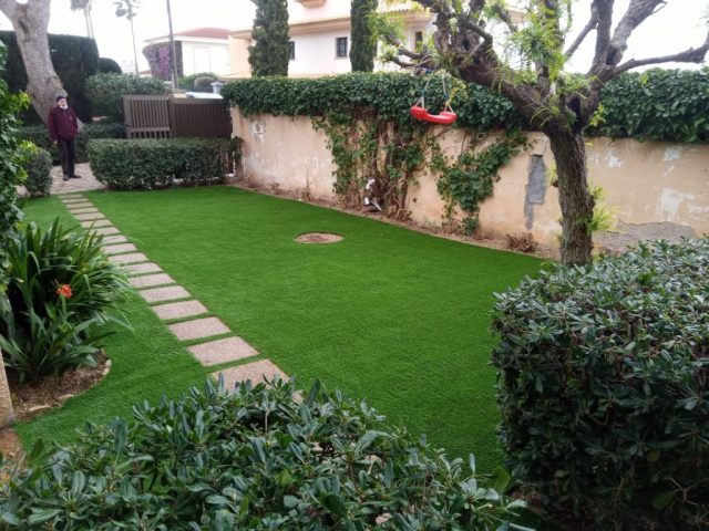 Jardín frontal con camino de losas y césped artificial alrededor de plantas y árboles